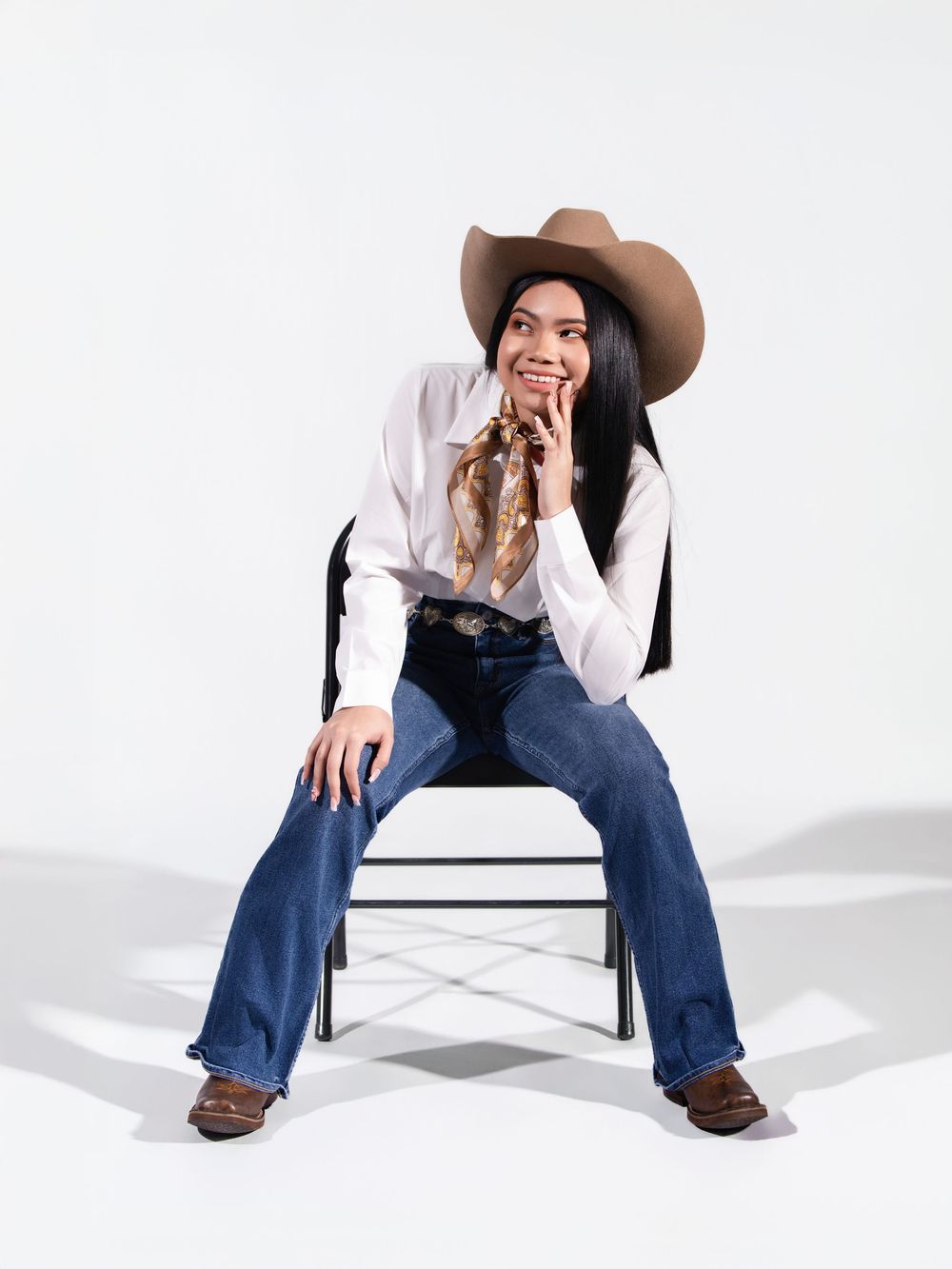 Woman in cowboy hat, white shirt, flare jeans, and boots sits on chair smiling in a western-inspired outfit.