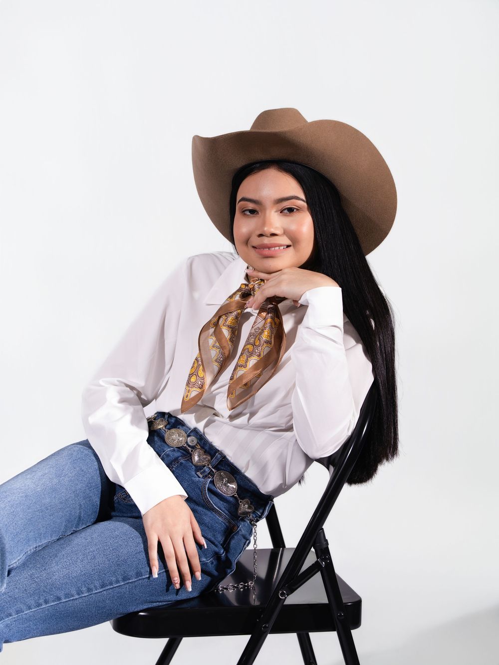 Smiling woman in cowboy hat, white shirt, scarf, and embroidered jeans seated on folding chair.