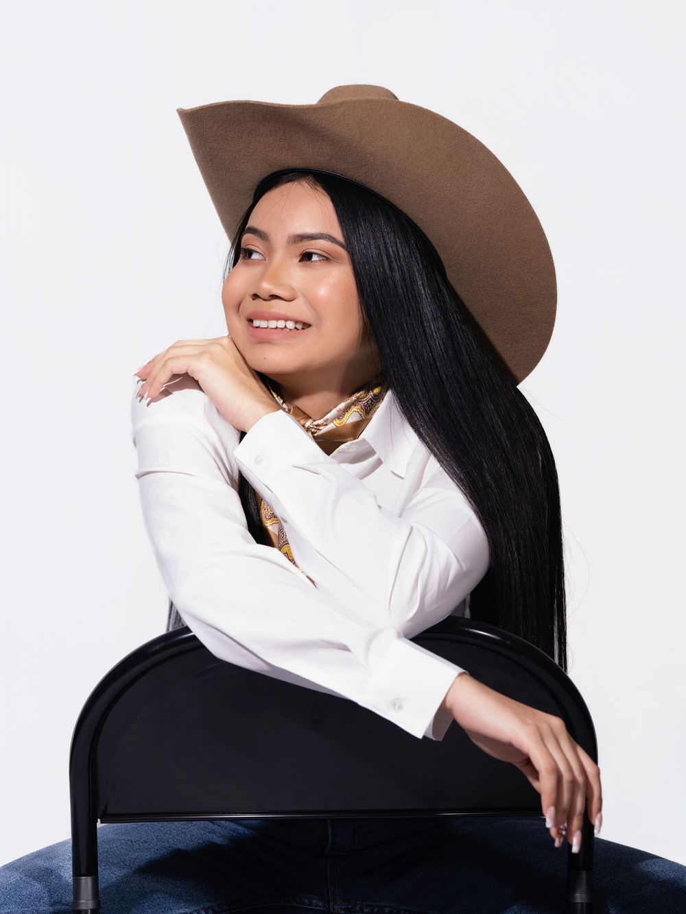 Young woman in a tan wide-brim cowboy hat and white shirt sits gracefully on a chair, smiling confidently.