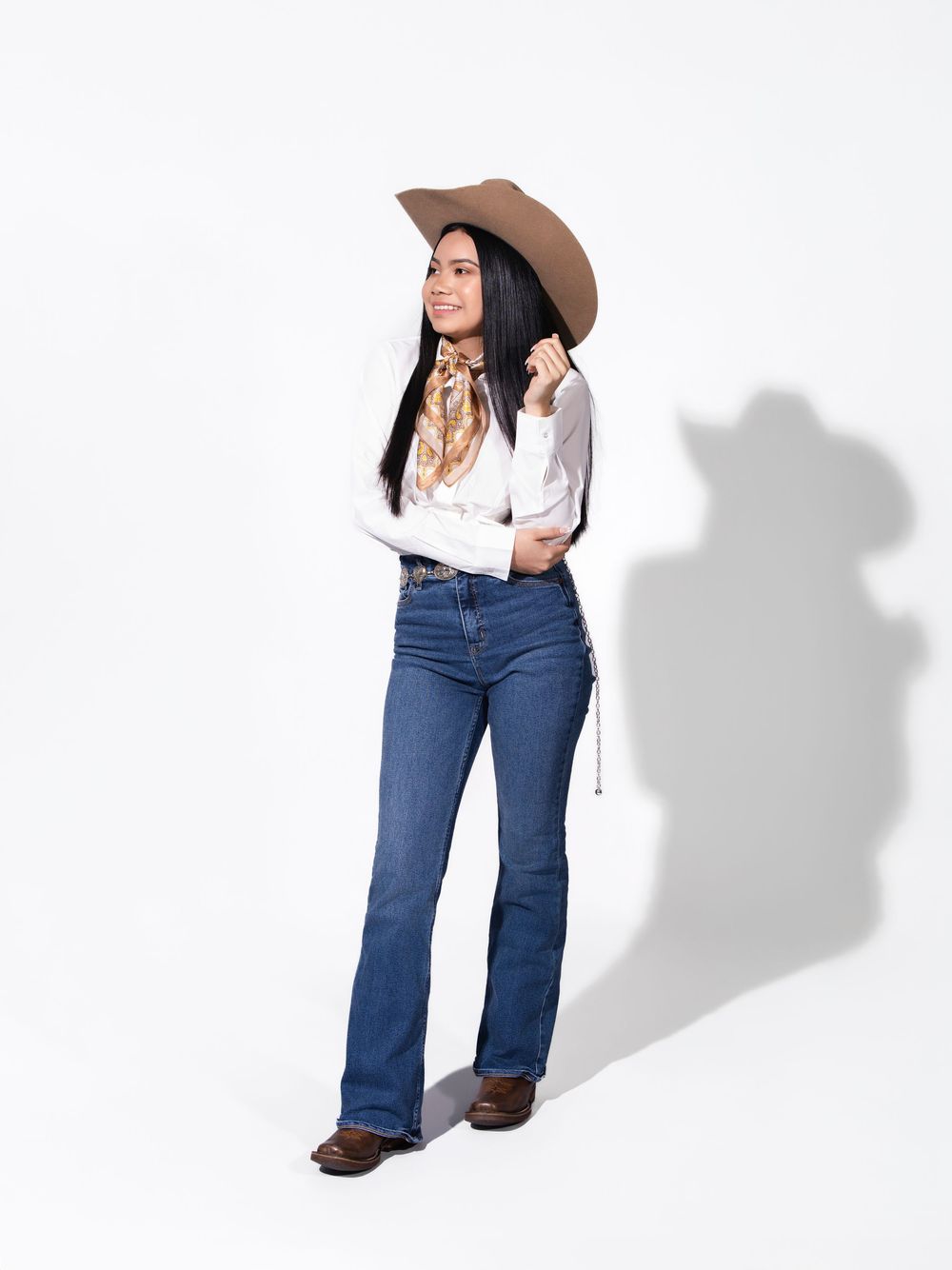 Woman wearing cowboy hat, white blouse, blue flare jeans, and brown boots poses against white background.