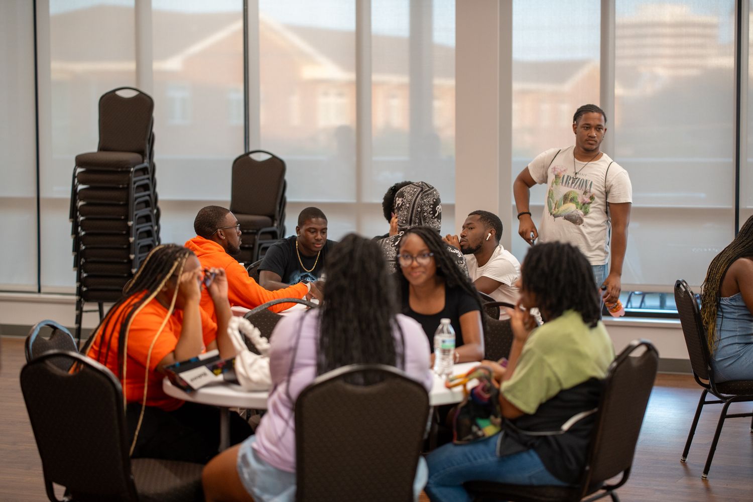 Groups of students sit at round tables in a bright, window-lined room, chatting while a man in a white shirt stands nearby.