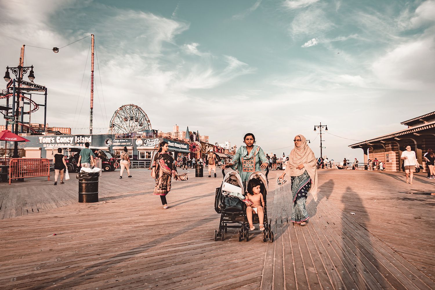 People walking on Coney Island boardwalk with ferris wheel and carnival rides in background.