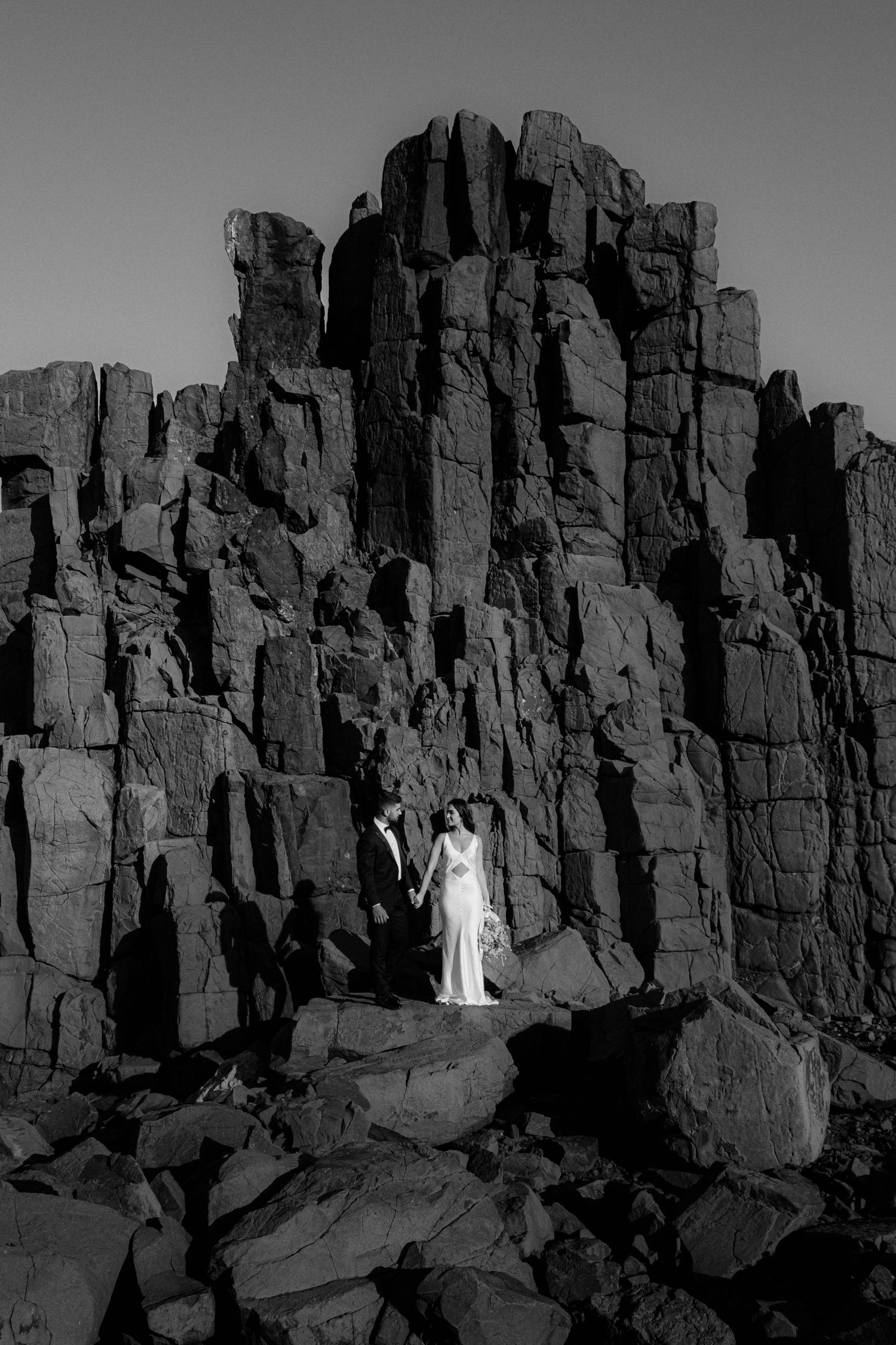 Wedding couple standing against dramatic black rock formation in monochrome.