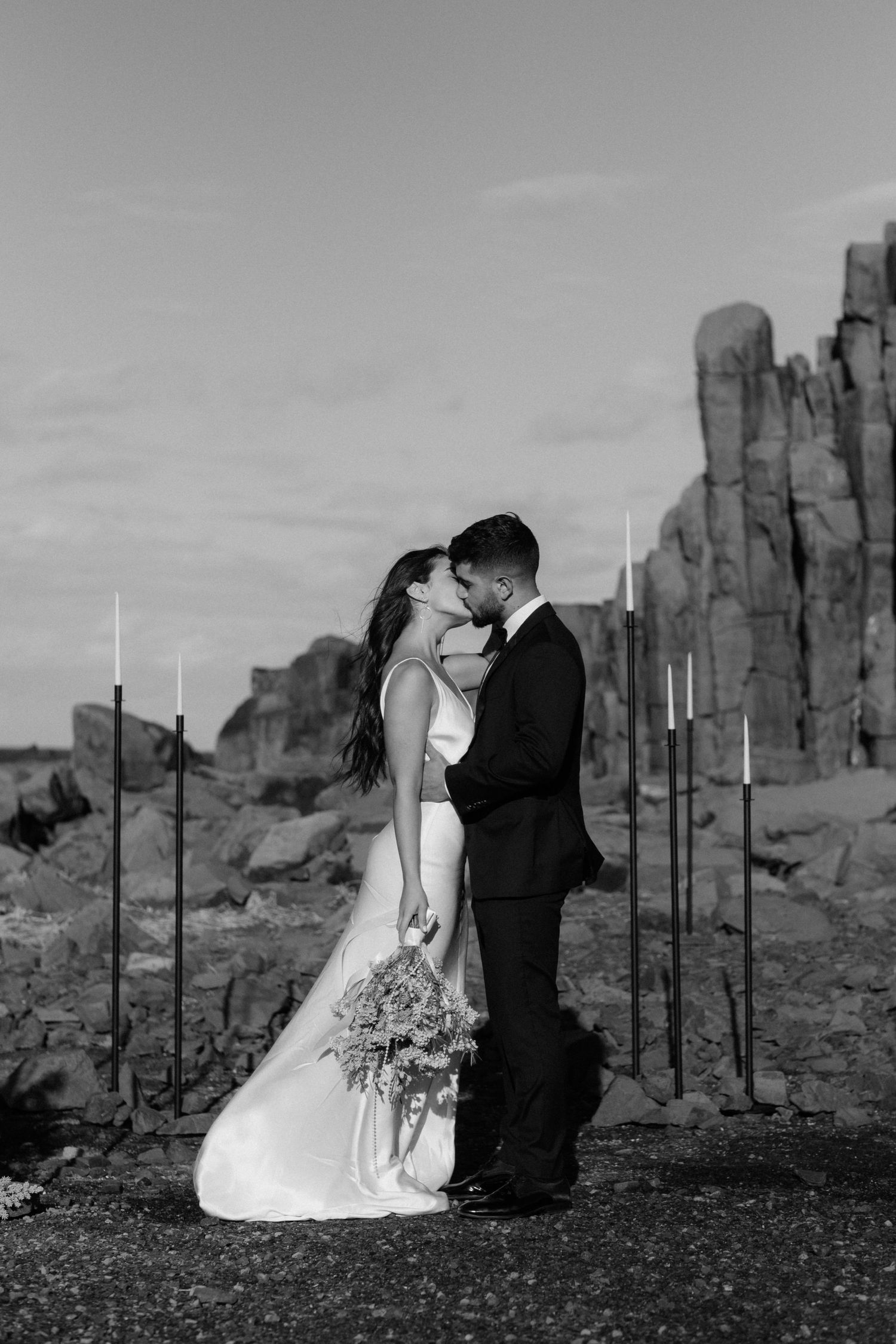 Black and white wedding portrait with tall rock formations and vertical poles in background.