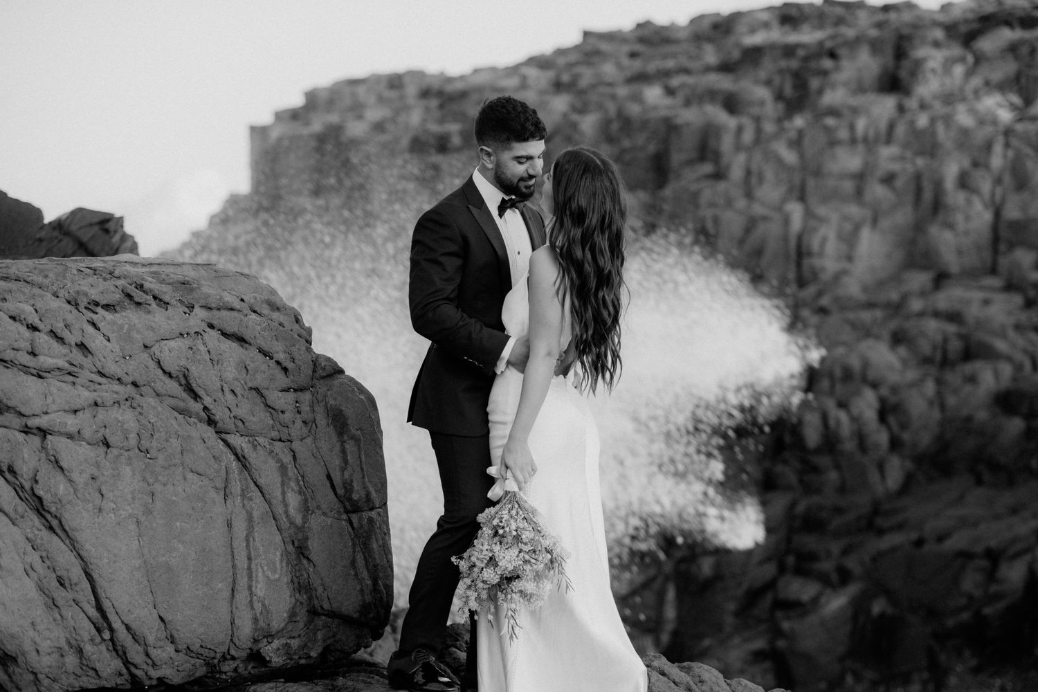 Black and white wedding portrait on dramatic desert cliff edge with wind-swept veil.