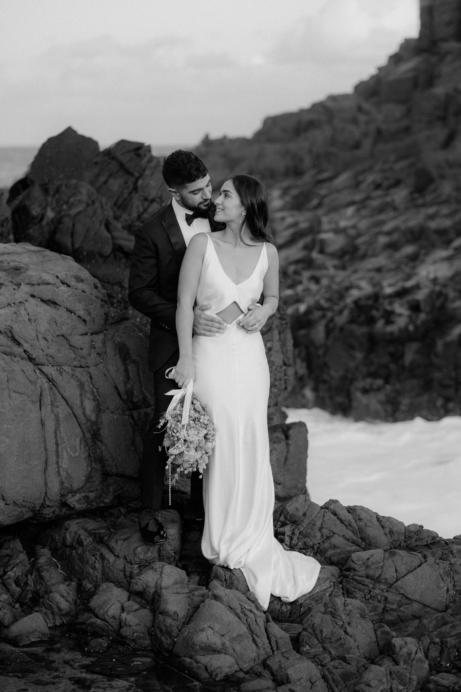 Wedding couple embracing on rocky coastal cliffs in elegant black and white photography.