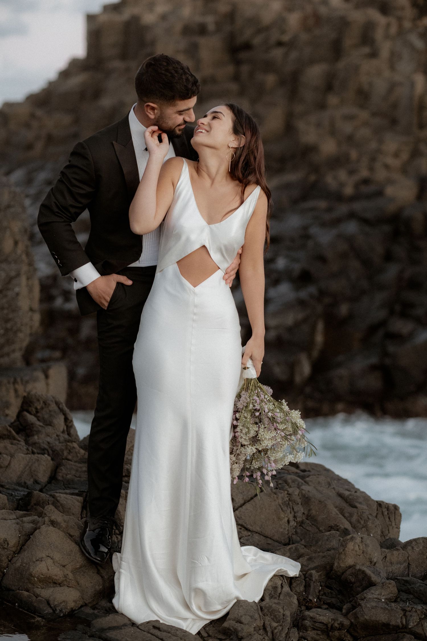 Elegant wedding photography on rocky coastline with crashing waves.