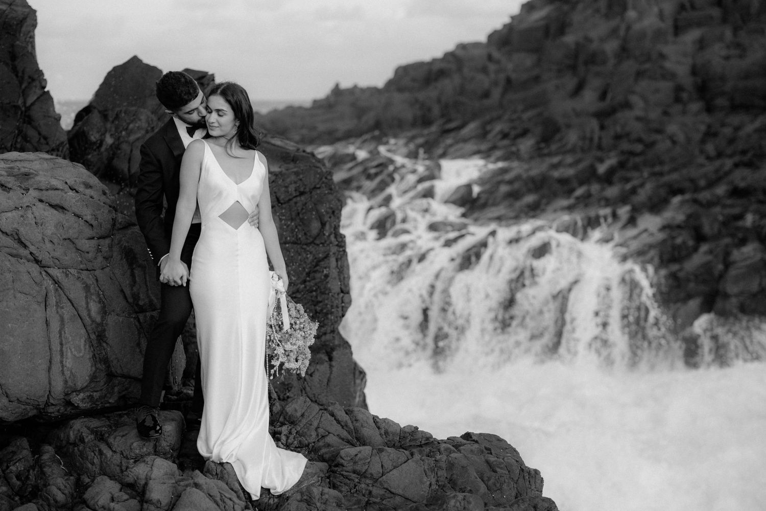 Black and white wedding portrait with dramatic waterfall and rocky cliffs in background.