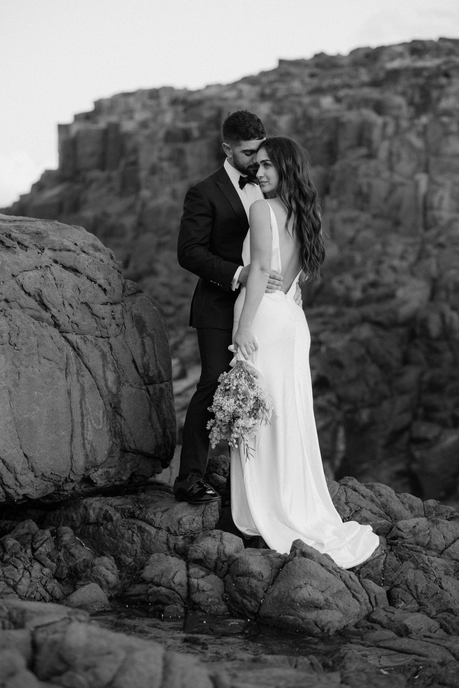 Romantic black and white wedding portrait against dramatic cliff backdrop.