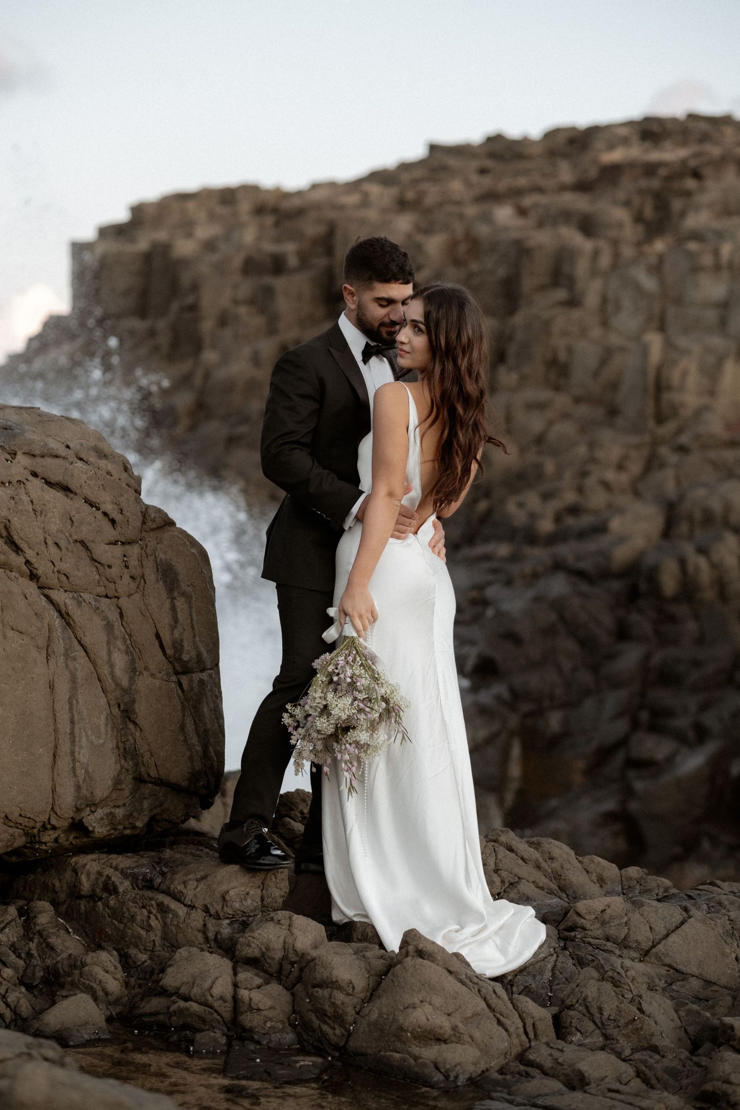 Wedding couple standing on rocky cliffs with dramatic coastal scenery.