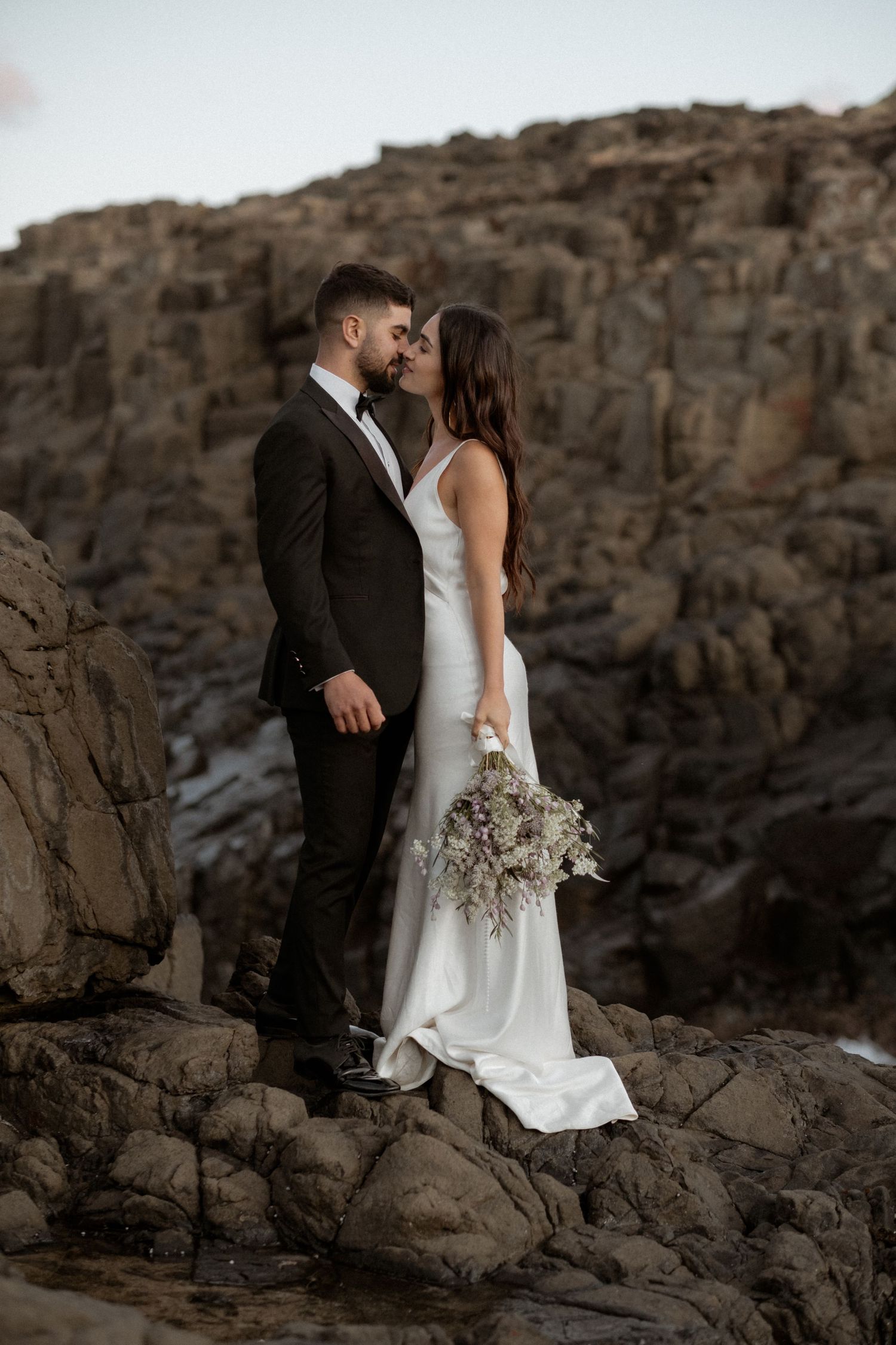 Intimate wedding moment captured on rugged coastal rocks at sunset.