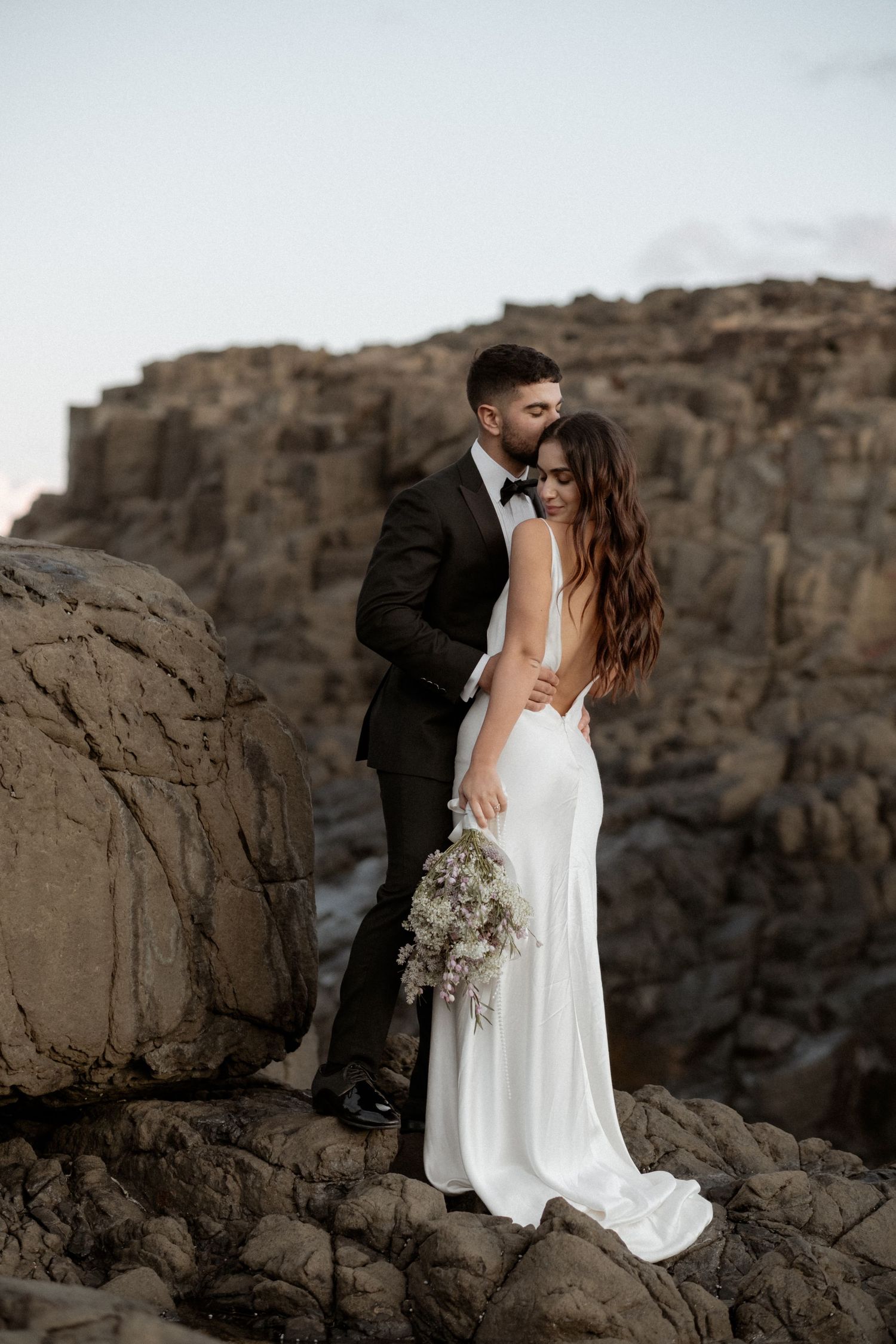 Elegant wedding portrait with dramatic rocky cliffs and ocean backdrop.