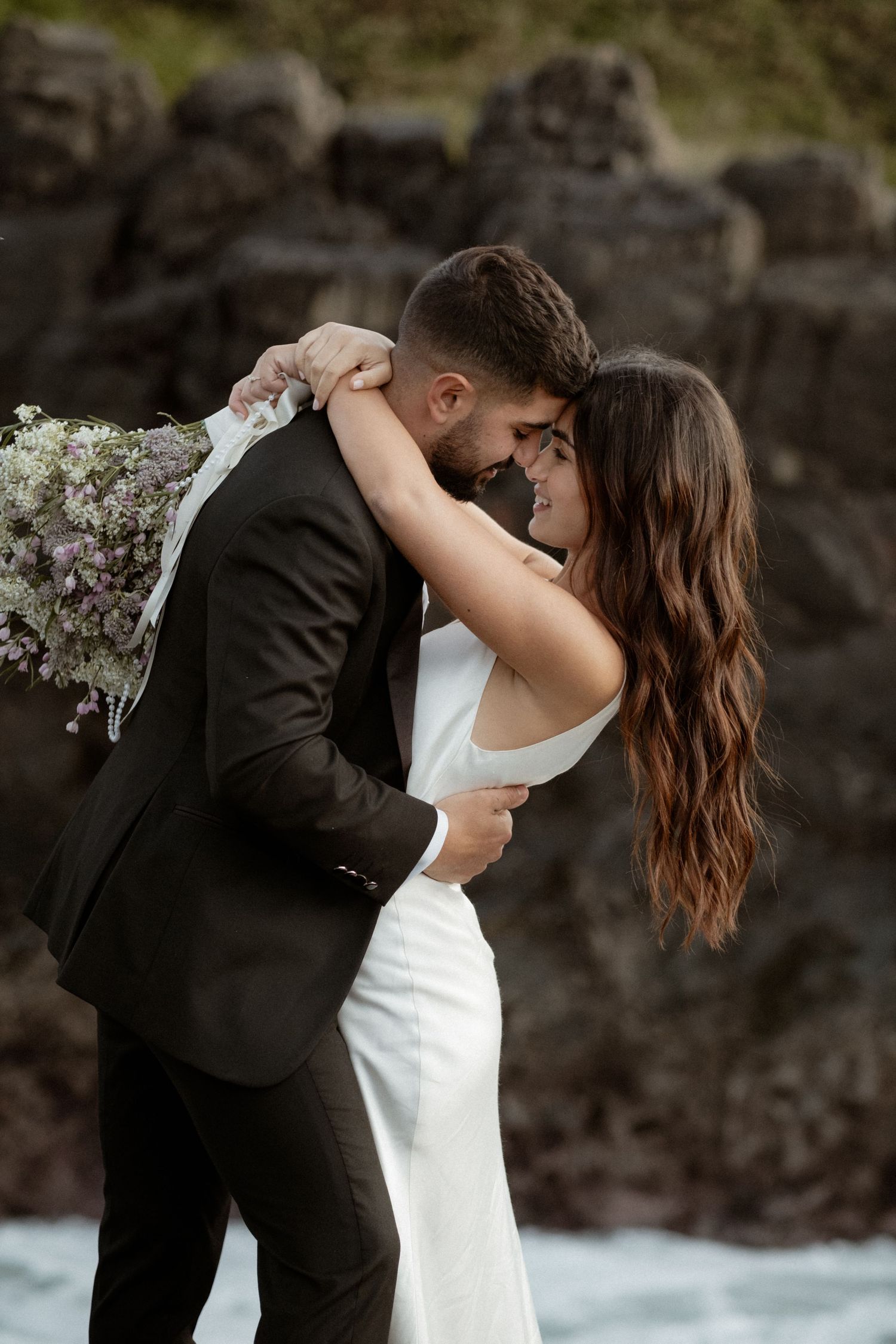 Romantic wedding couple embraces by rocky coastline with white bouquet and elegant attire.