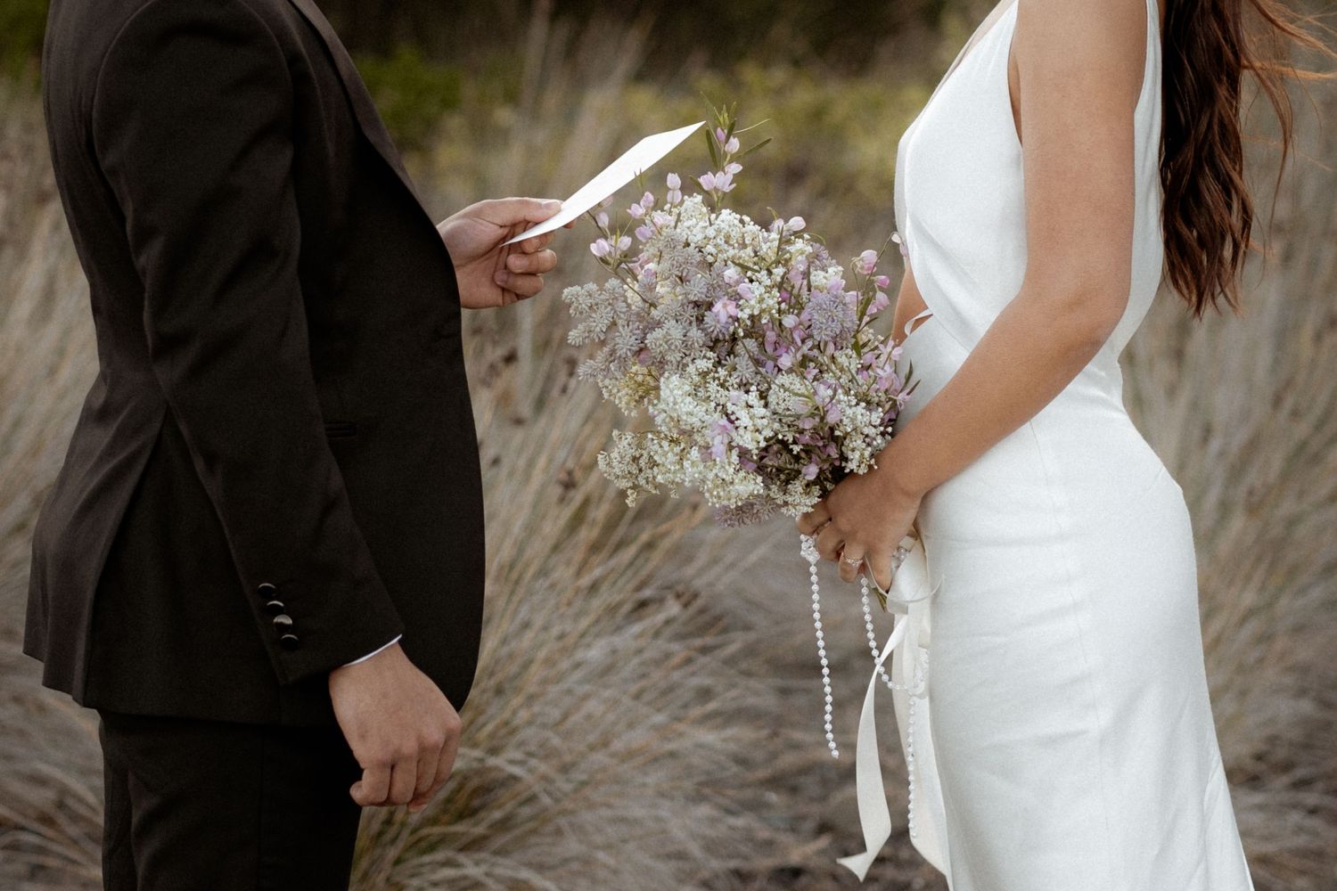 Elegant couple with white bouquet standing in coastal grass at wedding.
