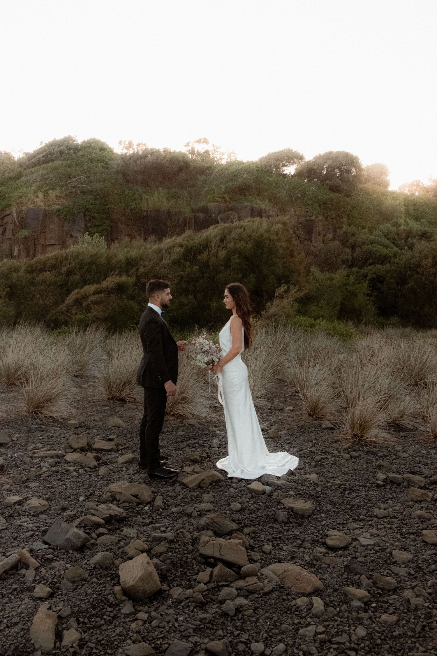 Wedding couple stands on rocky terrain at sunset in elegant attire.