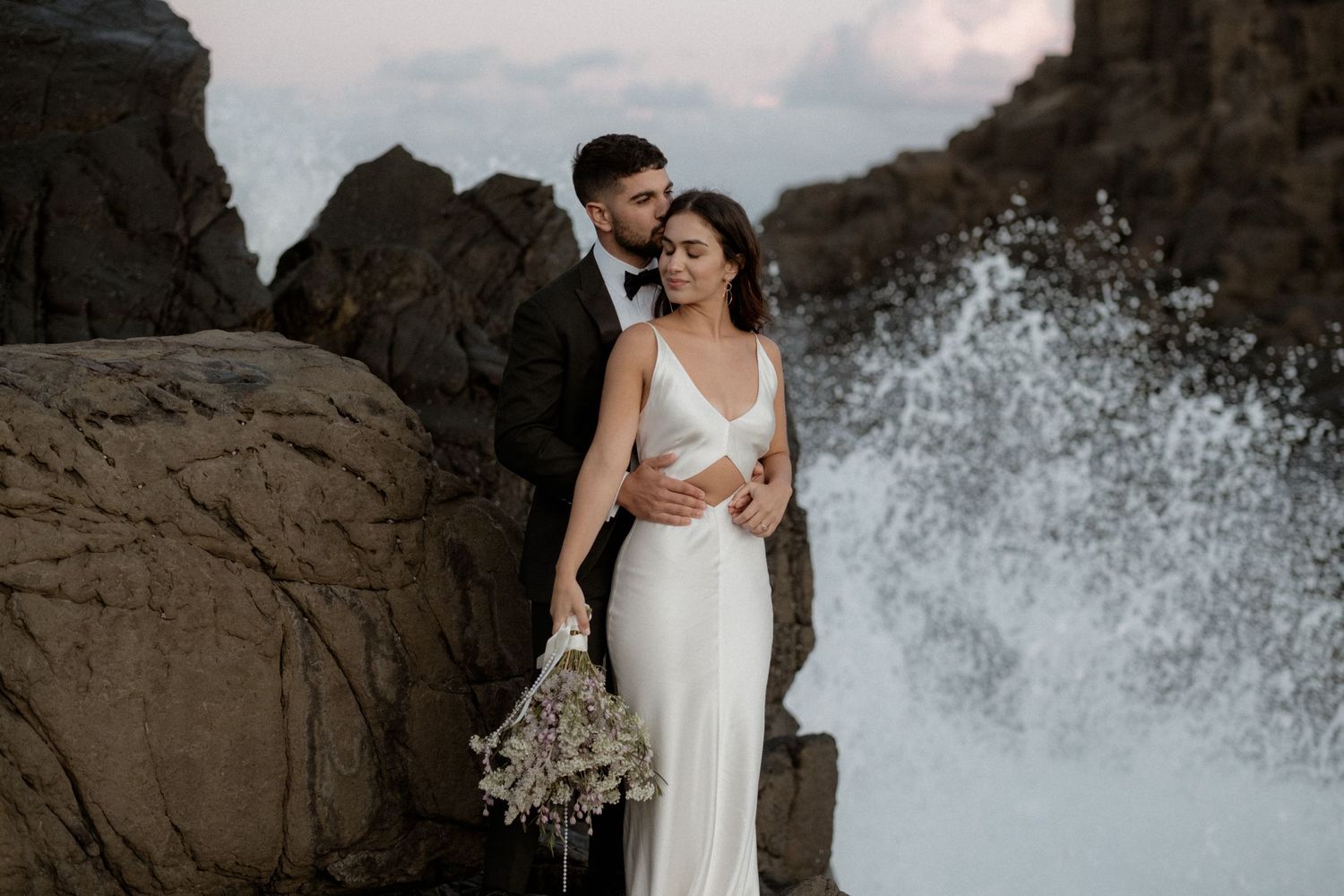 Dramatic wedding portrait with crashing waves against coastal rocks.