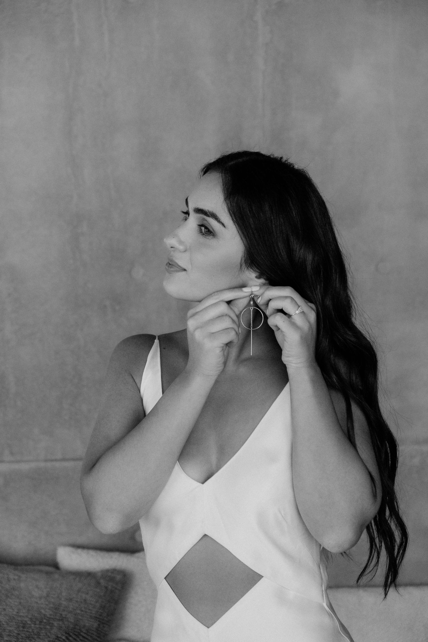 A woman in a white dress putting on earrings against a grey background in a black and white portrait.