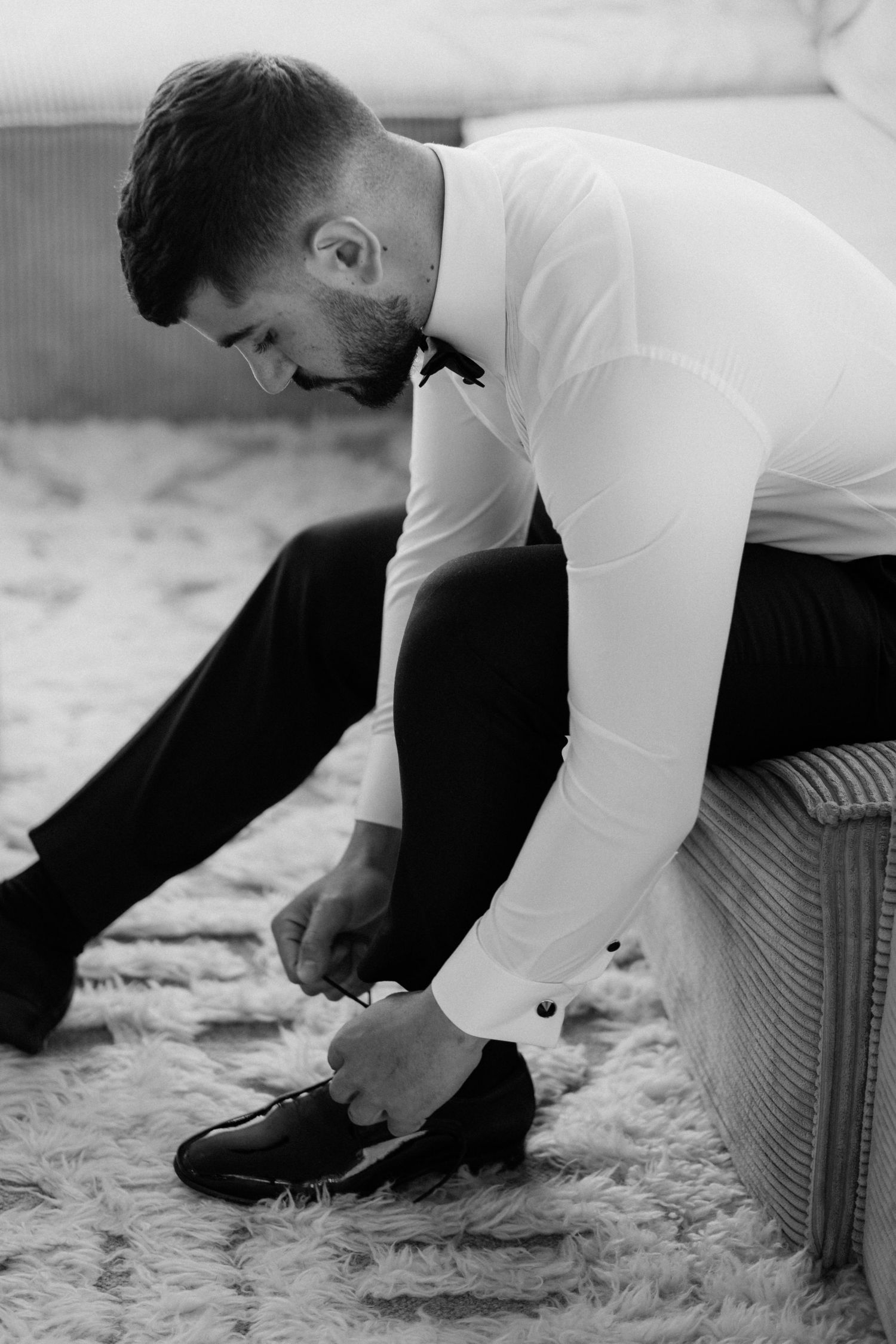 Man in formal white shirt tying his shoes while sitting on a plush carpet.