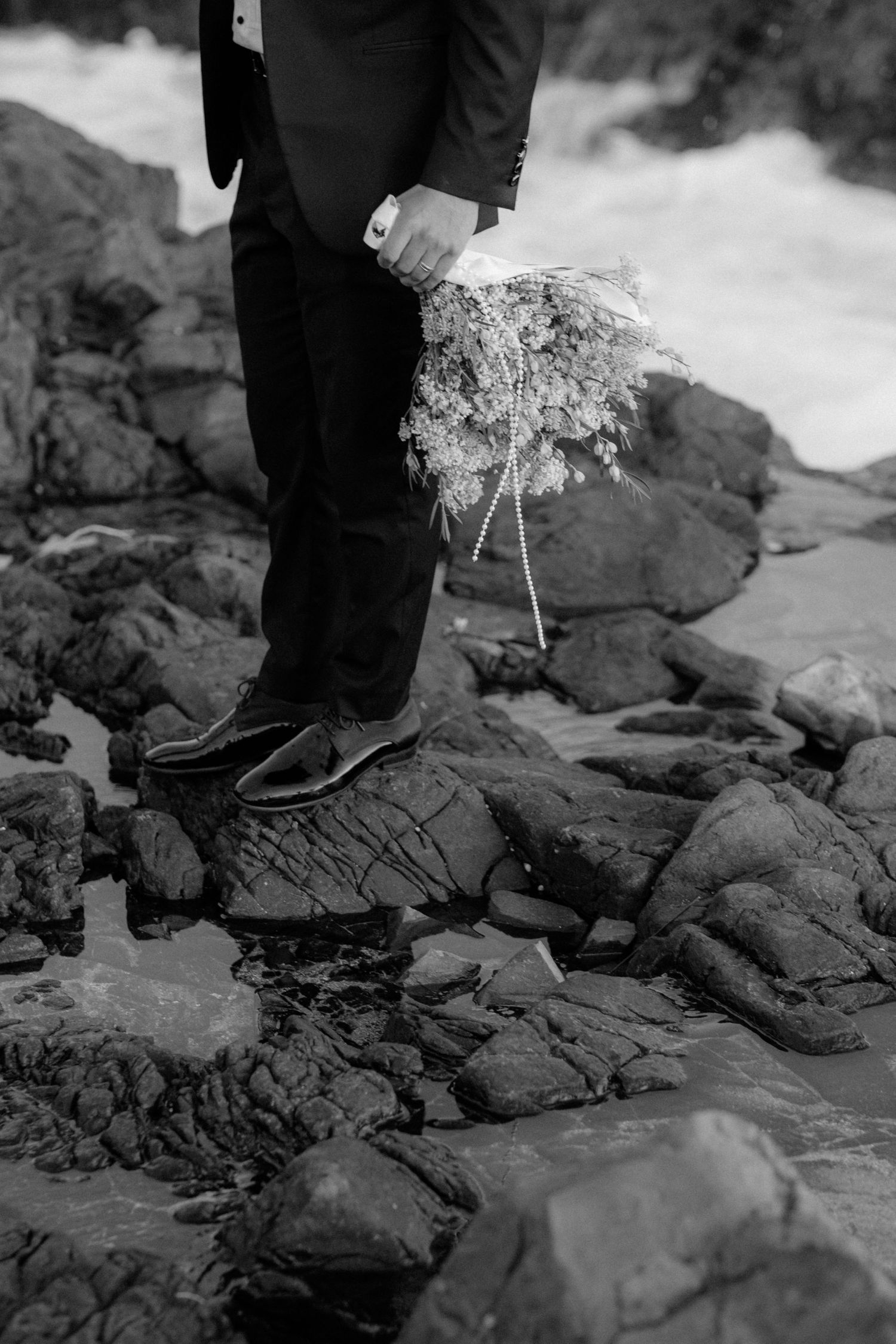 Black and white image of formal shoes and pant legs standing on rocky coastal terrain.