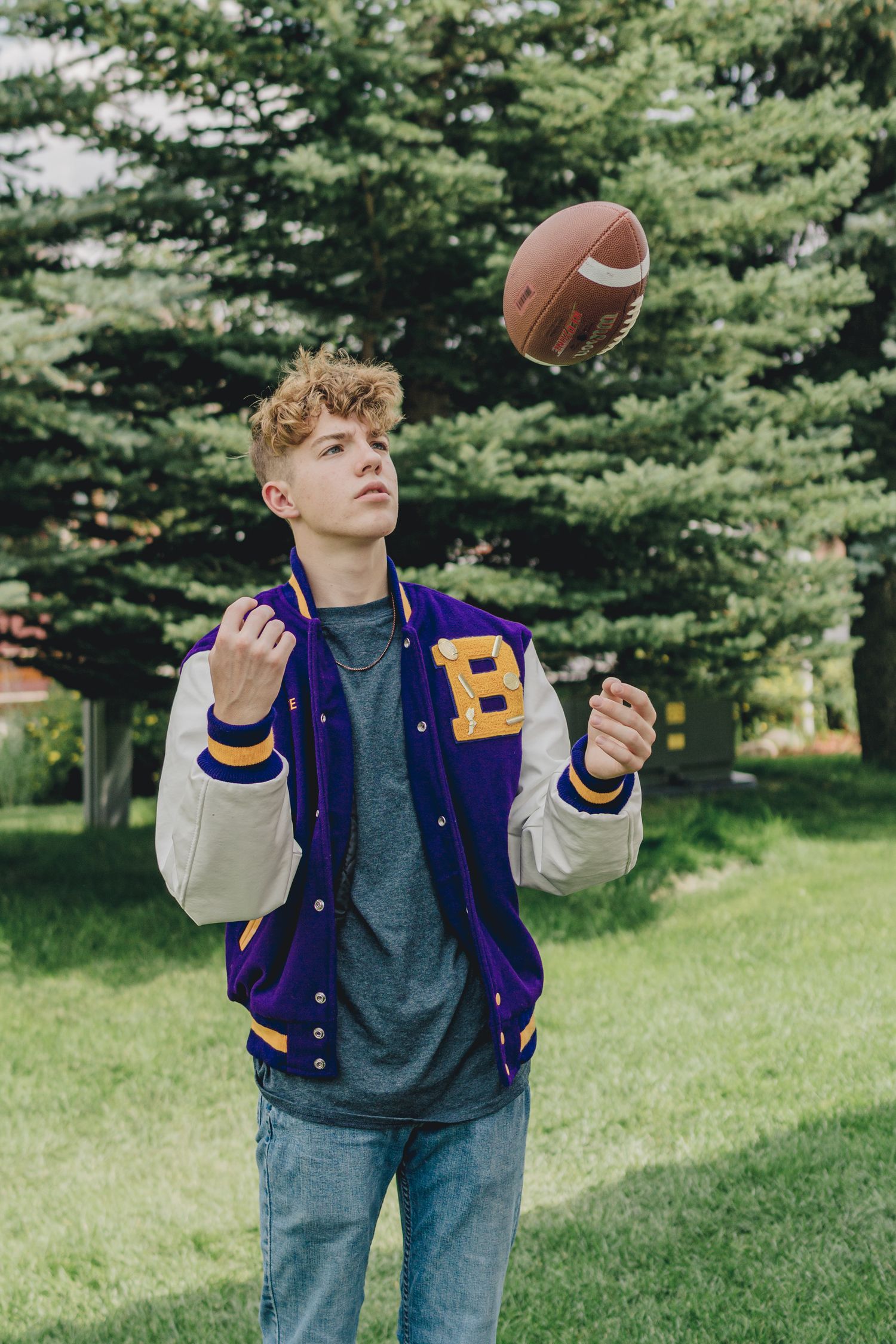 A teenage boy in a varsity jacket tosses a football in the air outdoors near tall trees.