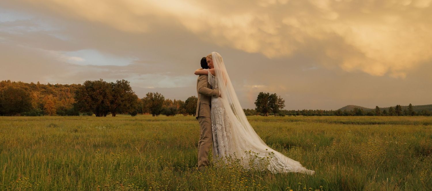 Bride and groom embrace in a lush green field at golden hour with dramatic clouds in the background.