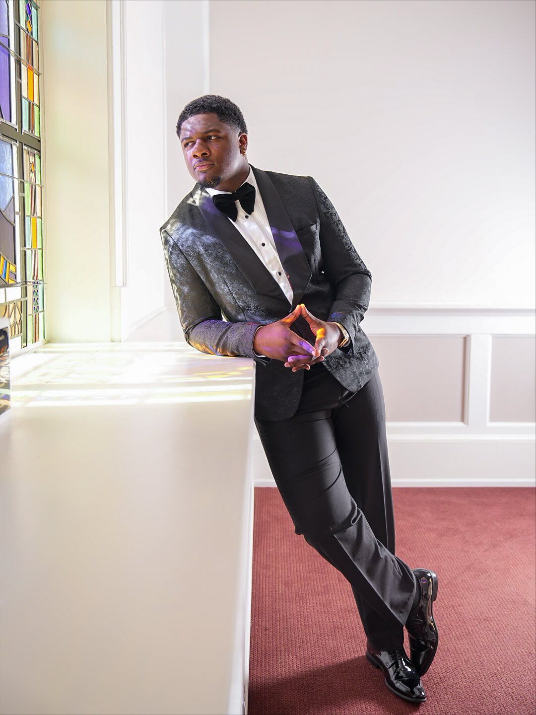 Man in a black patterned tuxedo and bow tie leans on a white ledge beside a stained glass window, gazing to the side.