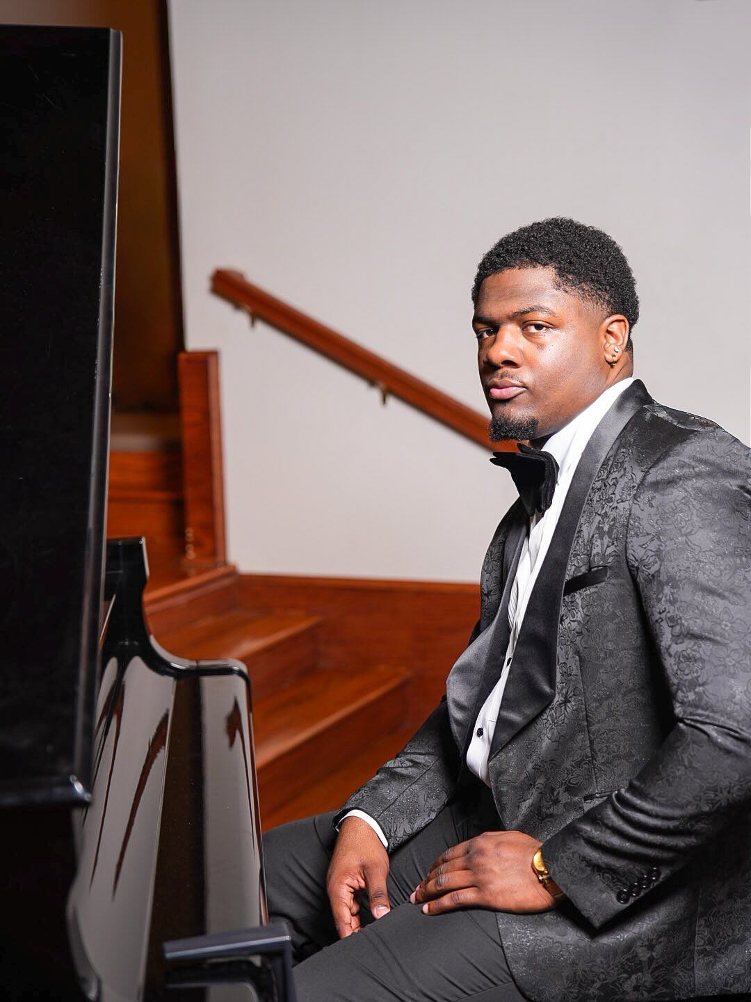 Man in a black jacquard tuxedo jacket and bow tie sits at a piano near a wooden staircase, looking toward the camera.