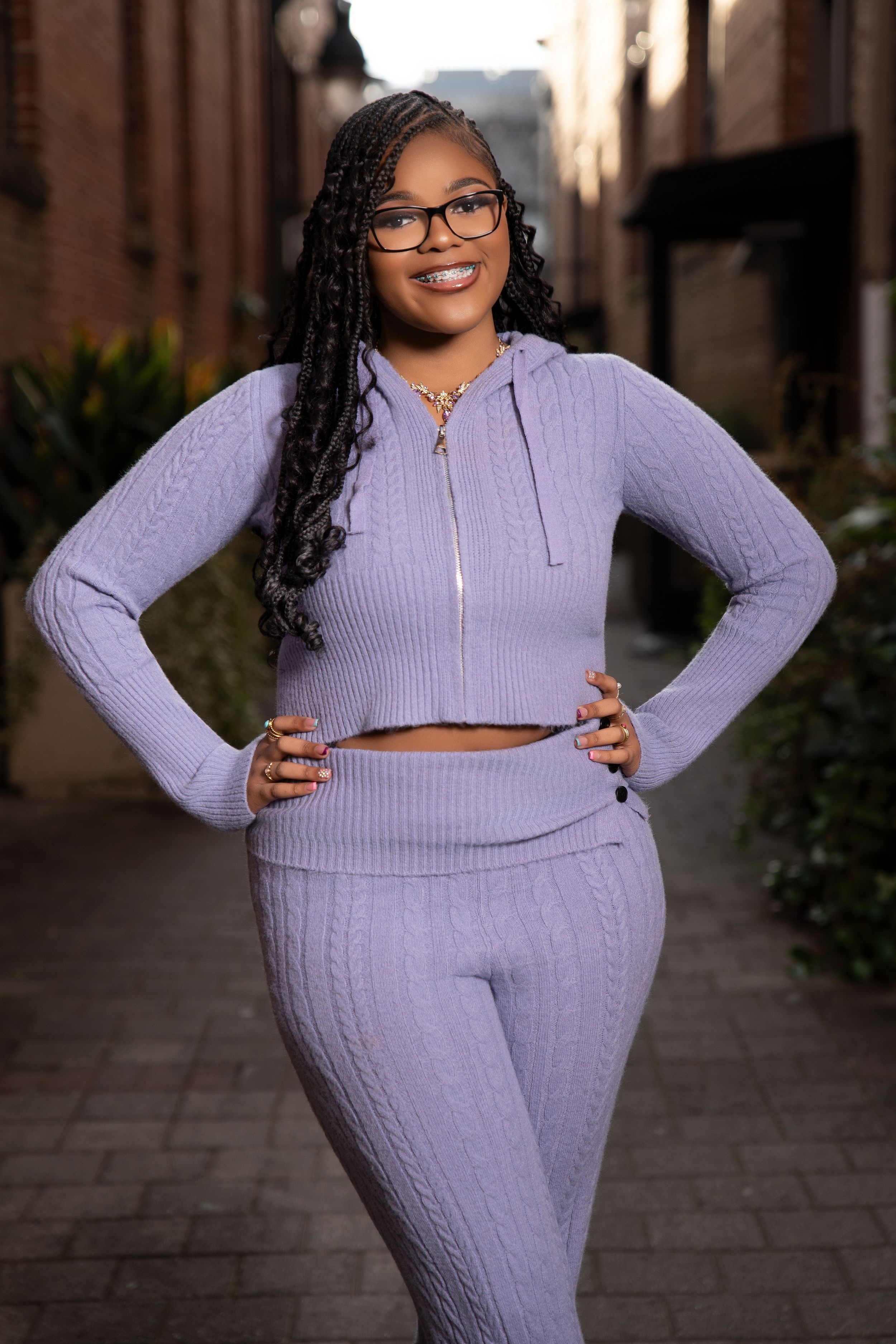 Smiling woman with long braids and glasses poses in a matching lavender cable-knit hoodie and pants on a brick-paved alley.