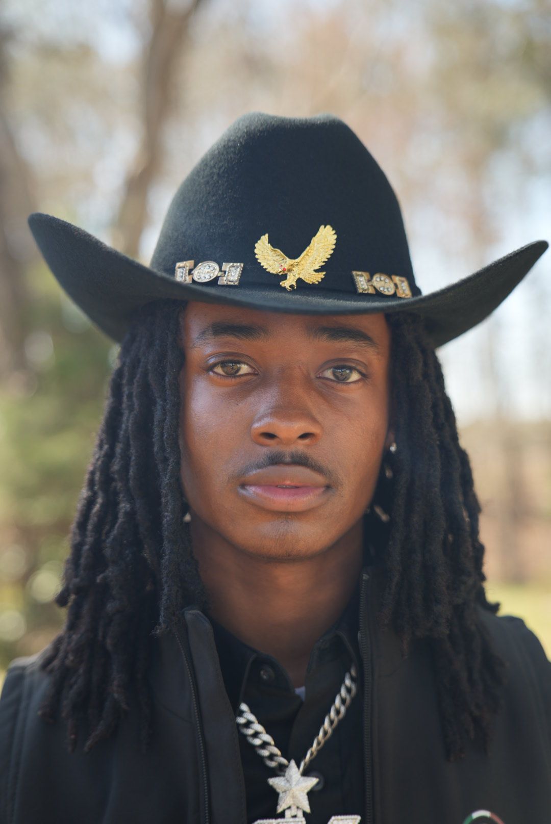 Young man with long locs wearing a black cowboy hat with a gold eagle pin and a silver star chain necklace outdoors.