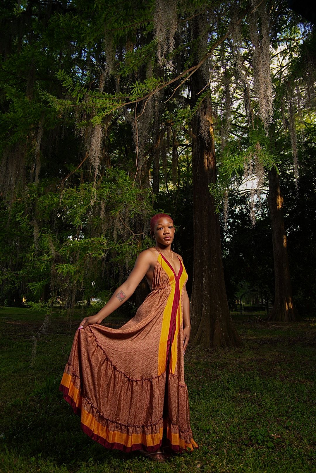 Woman in a flowing rust and gold maxi dress stands holding her skirt in a shaded park beneath tall trees draped with Spanish moss.