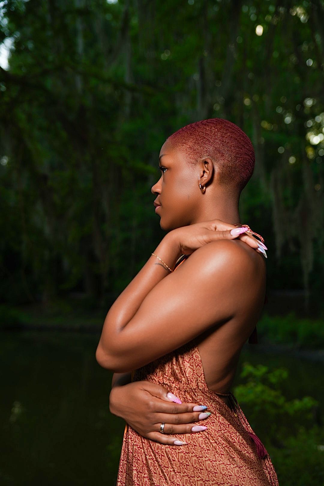 Profile view of a woman with short red hair and pink manicured nails embracing herself in a rust dress beside a dark, tree-lined pond.
