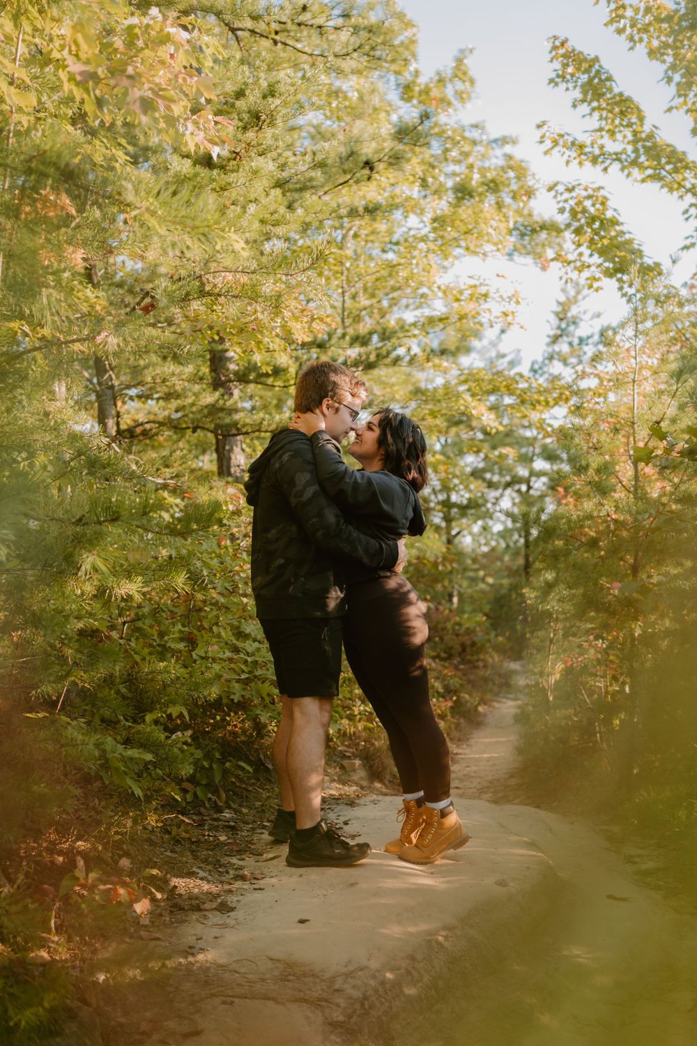 A couple stands on a forest trail, embracing and about to kiss amidst the greenery.