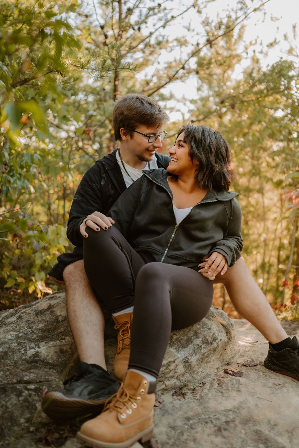 A couple sits on a rock in a forest, smiling and looking at each other affectionately.