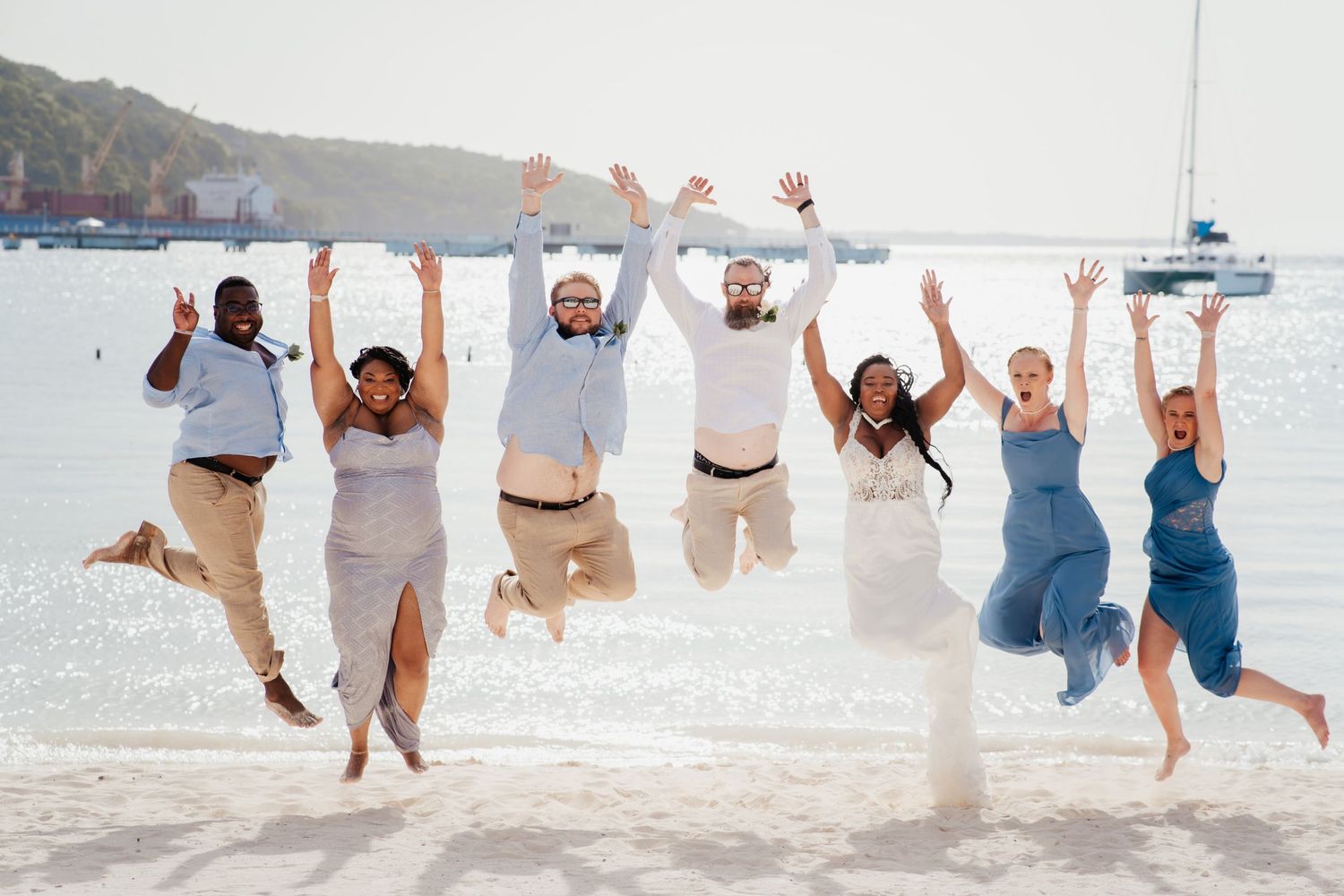 Wedding party jumping joyfully on beach with sailboat in background.