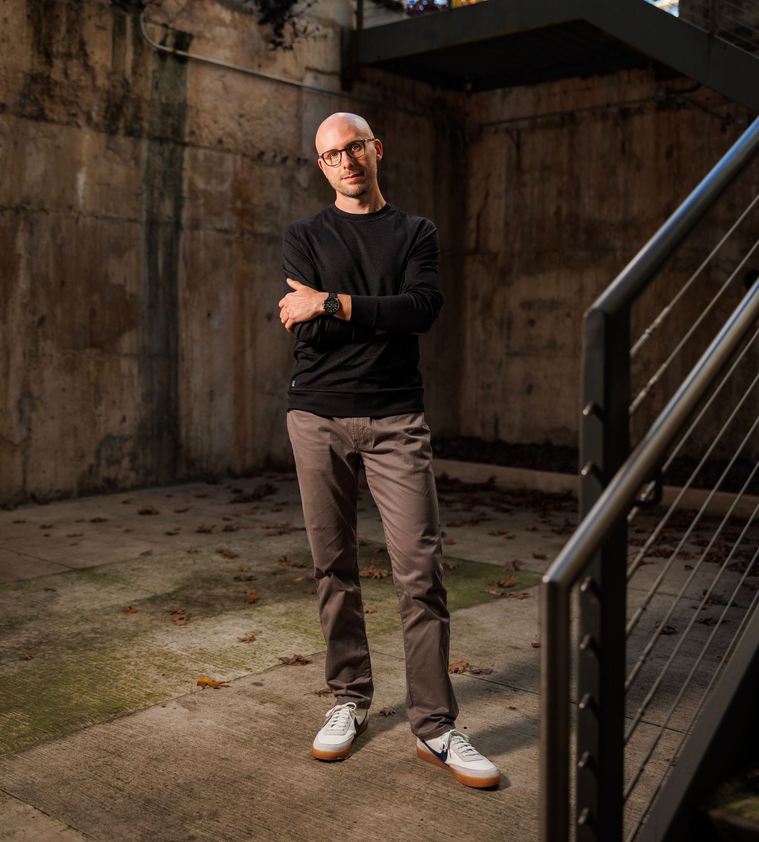 A person in a black sweater and khaki pants stands confidently in an industrial concrete space near metal stairs.