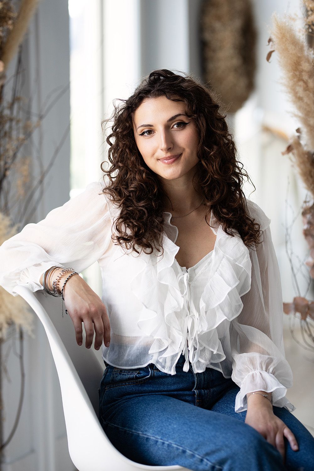 A person with curly dark hair wearing a white ruffled blouse sits casually in a modern white chair.