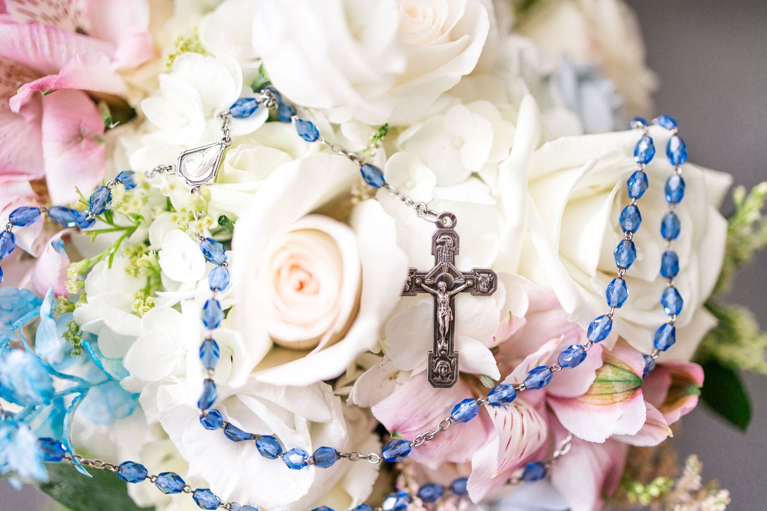 White roses and blue rosary beads with silver cross in a wedding bouquet arrangement.