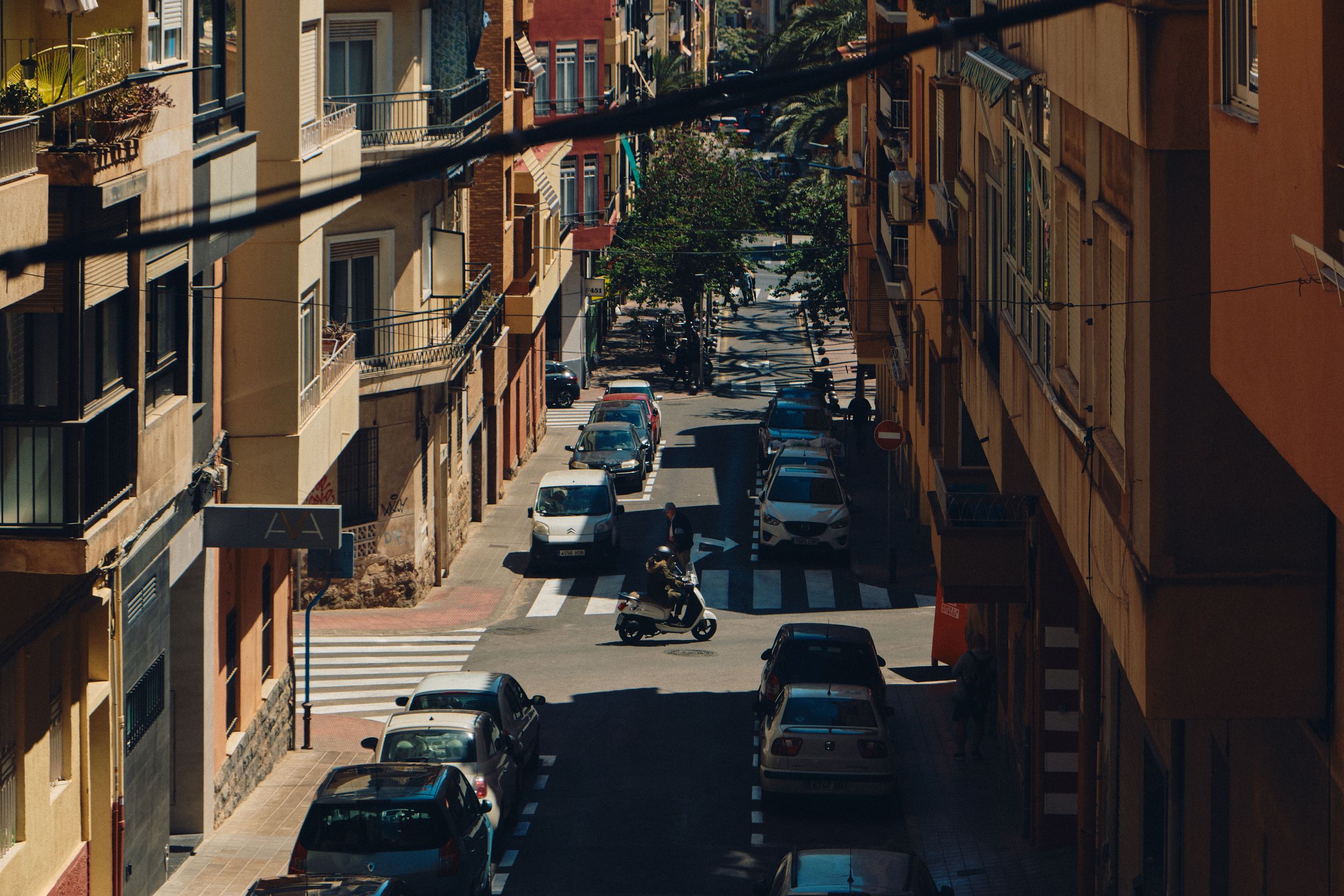 Narrow street lined with parked cars between residential buildings.