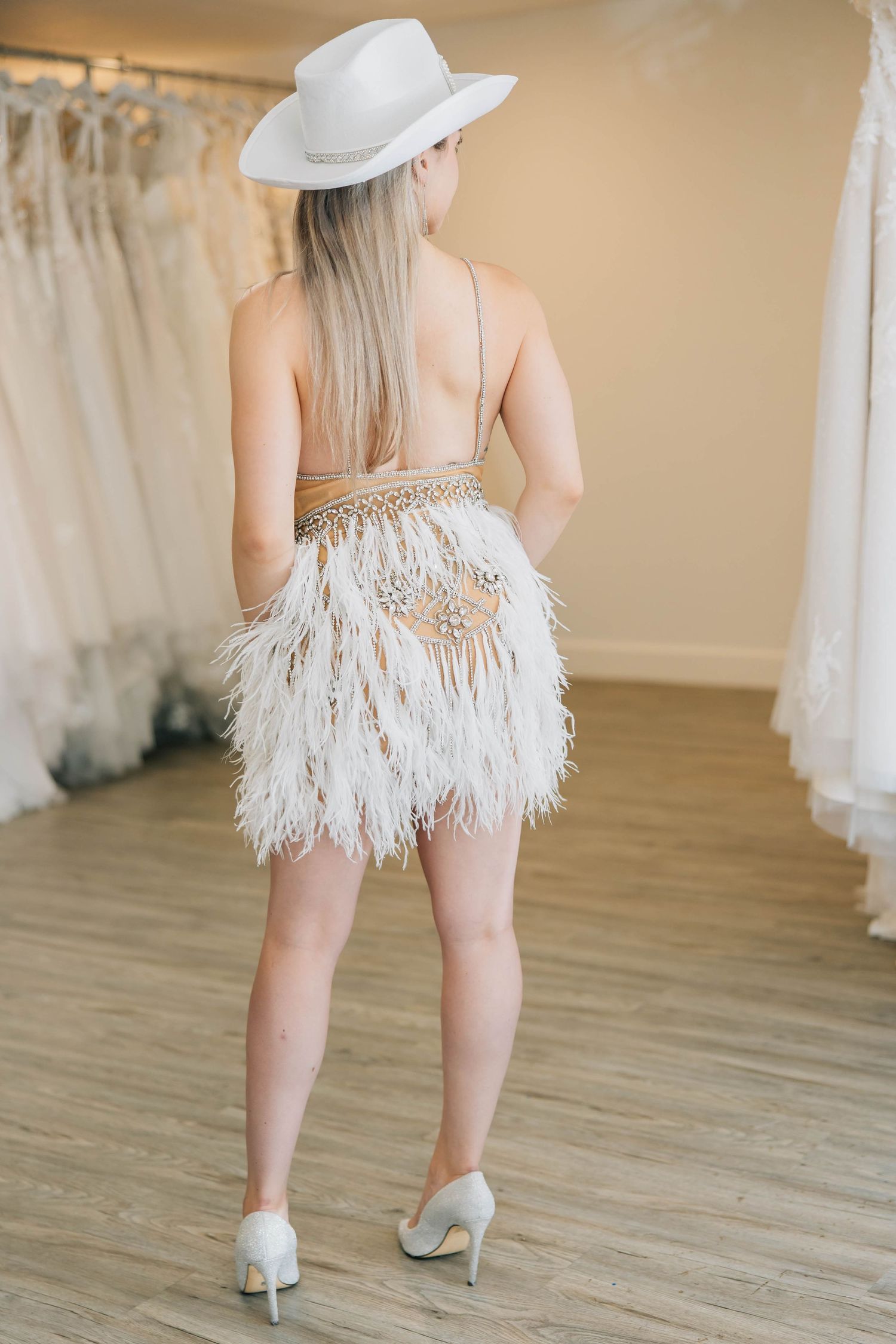 Woman viewed from behind wearing white feather mini dress and white cowboy hat in bridal shop.