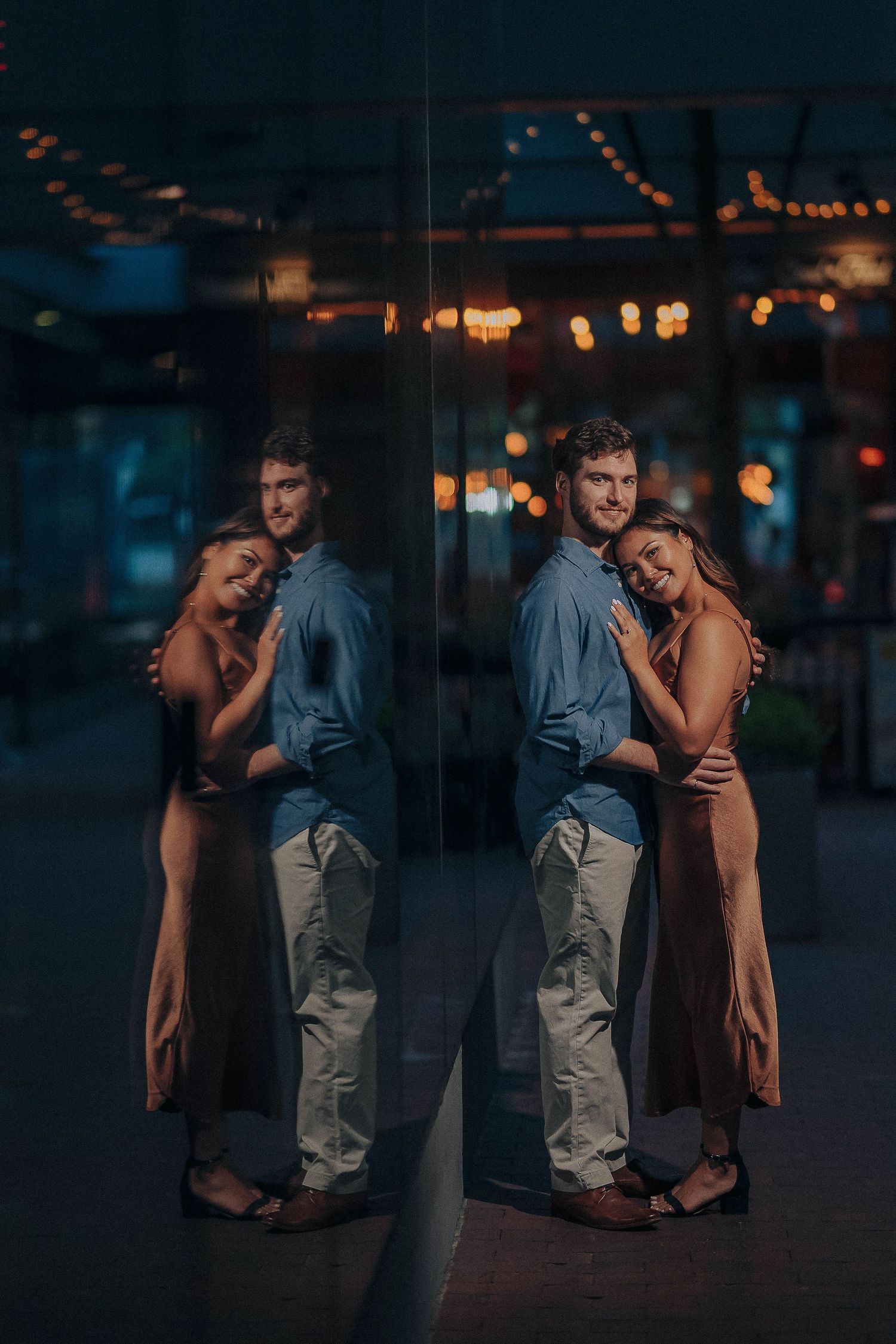 Romantic nighttime photo with string lights and window reflections showing a couple embracing in moody lighting.