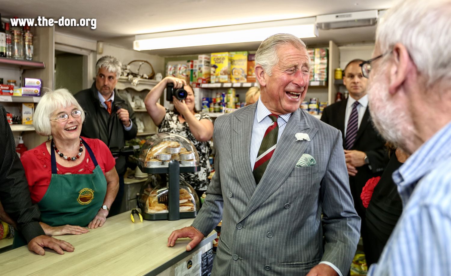 King Charles laughing with staff at a traditional deli shop counter during a formal visit to North Wales