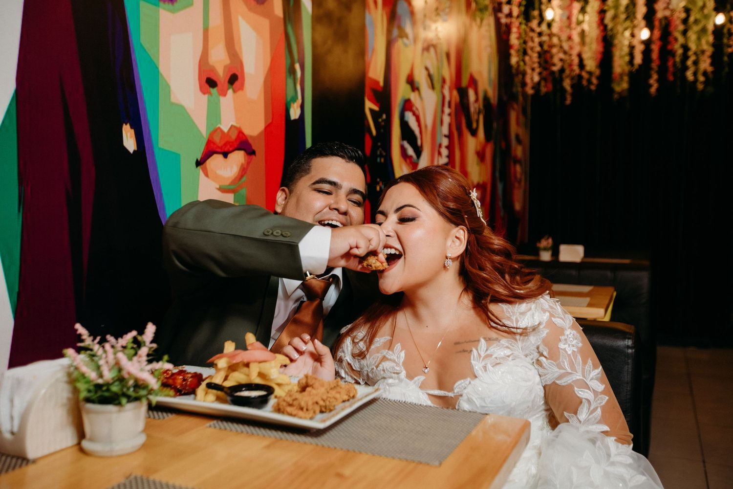 Couple sharing food and laughing during wedding reception in dimly lit venue with decorative lighting.
