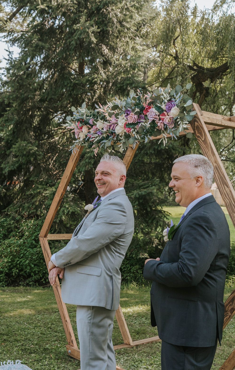 Two people in formal attire share a lighthearted moment before a wooden arch at an outdoor wedding.