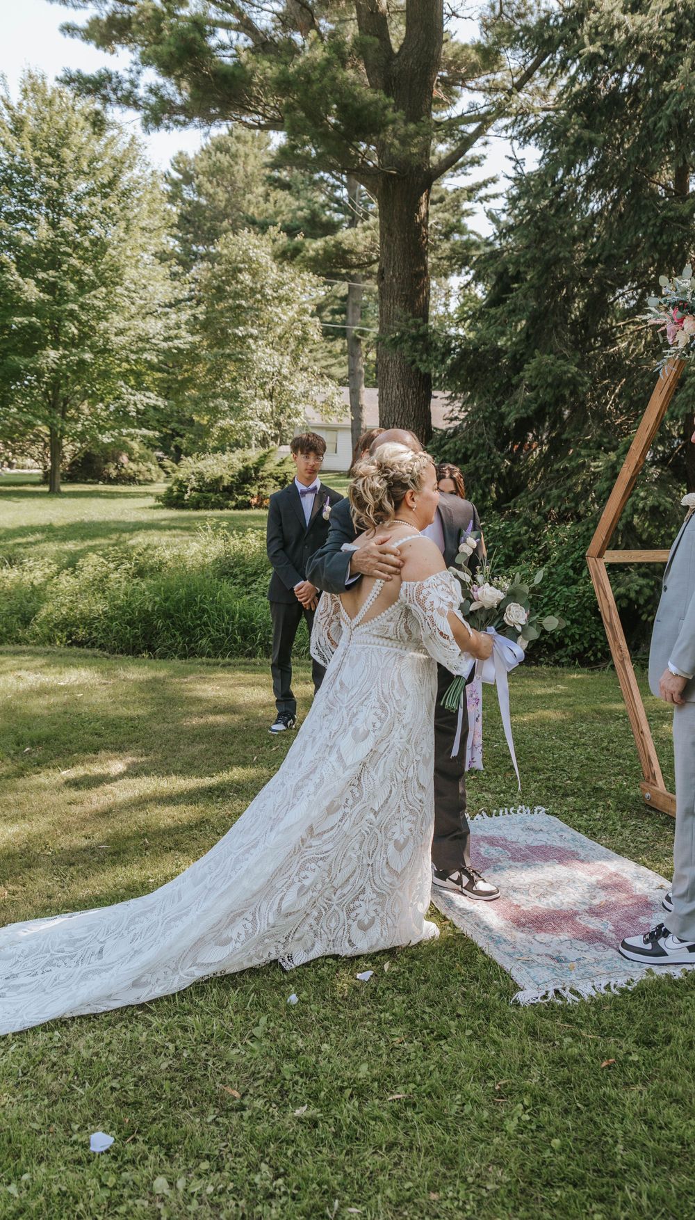 An outdoor wedding ceremony under trees with a wooden hexagonal arch and floral decorations.