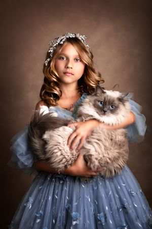 Young child in a blue tulle dress adorned with butterflies holding a large Ragdoll cat against brown backdrop.