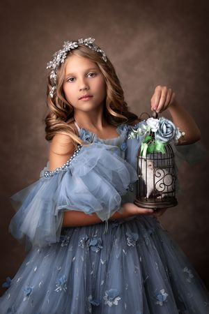 Child wearing a flowing dusty blue princess dress holds a decorative bird cage with blue flowers in ethereal portrait setting.