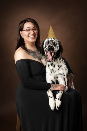 A spotted Dalmatian dog wearing a gold party hat yawns while sitting with someone in a black dress against a brown backdrop.