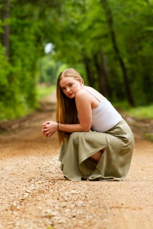 A person in a white tank top and olive skirt crouches on a dirt path surrounded by lush green trees.