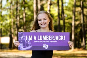 Person holding a purple banner with text 'I'M A LUMBERJACK!' against a sunny forest background.