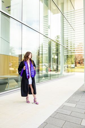 Young woman in graduation gown and purple stole poses confidently outside a modern glass building.